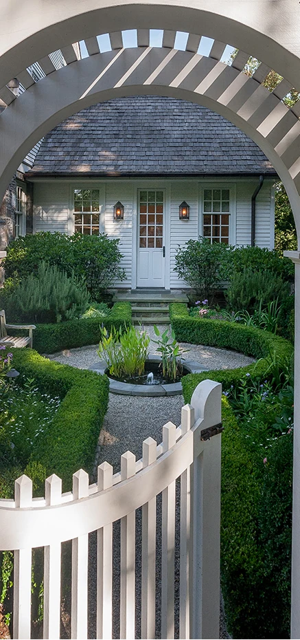 A white lattice arbour arch and picket gate framing a small formal parterre with clipped hedging, a gravel path and a circular water basin.