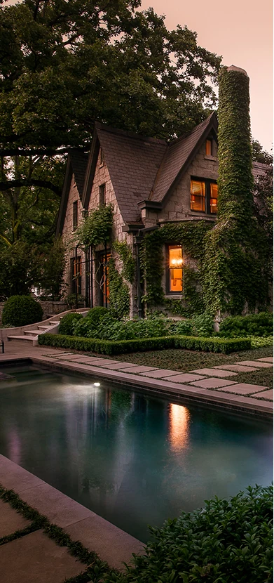 An ivy-clad stone house glowing from within at dusk, reflected in a still rectangular pool edged with stone paving and low clipped hedging.