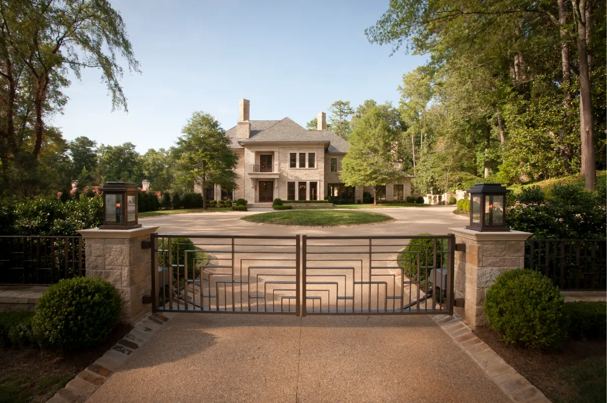 A bronze entry gate between stone piers with lanterns, opening onto a circular gravel motor court and a limestone house framed by boxwood and mature pines.