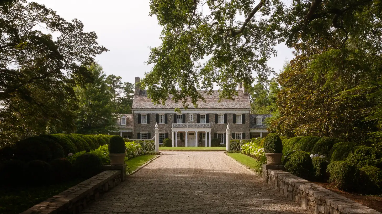A stone Georgian house at the end of a pea-gravel drive, flanked by deep clipped hedges and hydrangeas beneath a mature hardwood canopy.