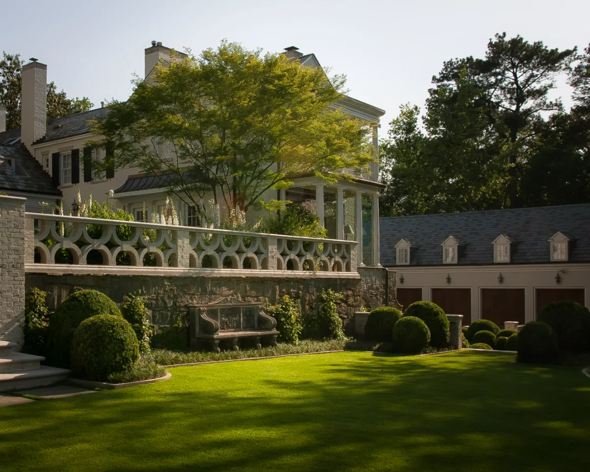 A level lawn edged with clipped boxwood domes below a stone terrace with a pierced limestone balustrade, foxgloves in flower and a Japanese maple against the house.